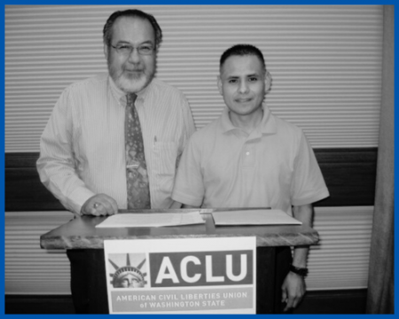 A black and white photo of two men standing behind a podium with an ACLU-WA sign on it