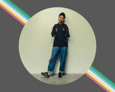 A self portrait of Jordan set on top of a muted black background and group of this diagnal stripes in the colors of the disability pride flag. 