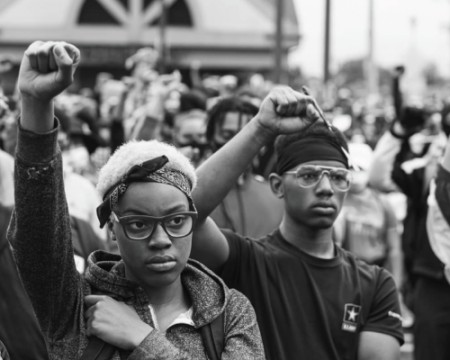 Two Black individuals stand in a crowd of demonstrators with their hands raised in a fist.