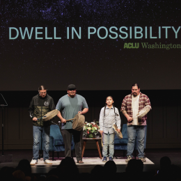 Four people stand on stage playing hand drums in front of screen projecting Dwell In Possibility 