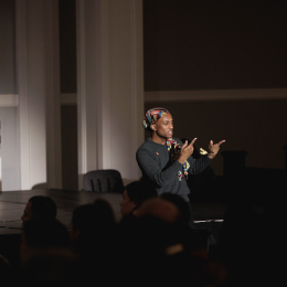 A person wearing a colorful headscarf standing in a spotlight and signing to an audience 