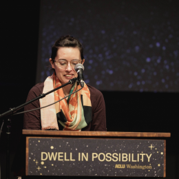 A person wearing glasses and a colorful scarf looking down while sitting behind a podium