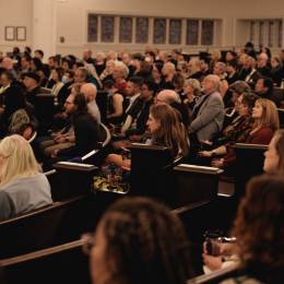 A room filled with rows of people sitting in pews facing a stage 