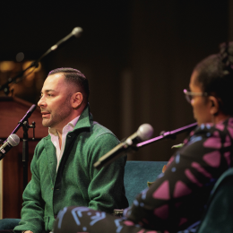 Two people talking into microphone stands one in the foreground and one in the background 