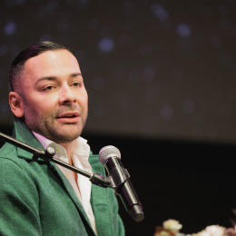 A speaker wearing a white shirt and green jacket speaking into a microphone