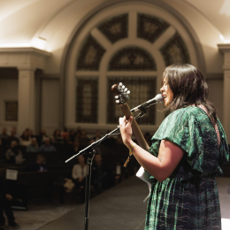 A musician wearing a green dress playing guitar and singing into a microphone 